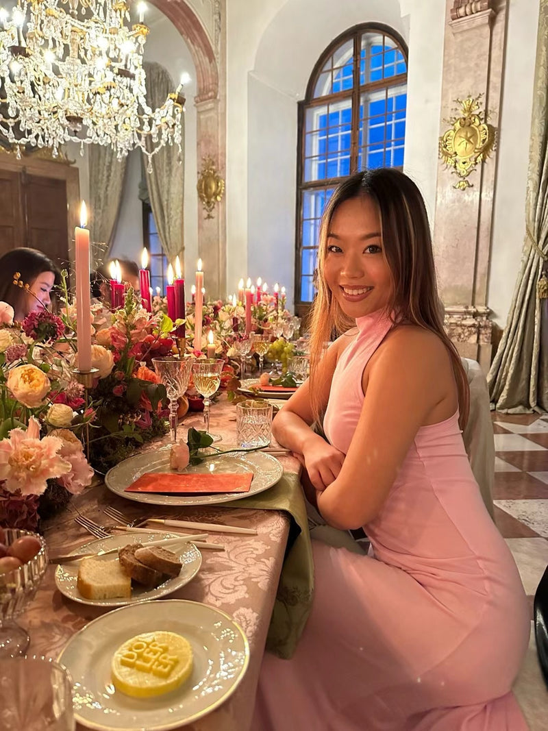 Woman in a pink dress sitting at a elegantly set dinner table with candles and flowers.